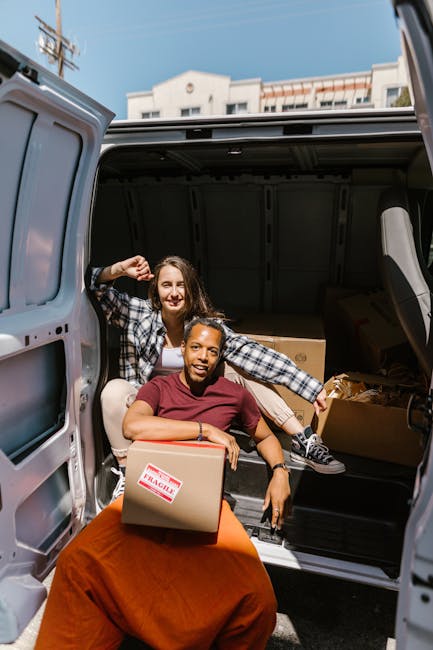 Two women are engaging in a house removal process at the rear of a grey cargo van, which is parked outdoors in a residential area. The woman sitting at the edge of the van’s loading area is holding a cardboard box labeled 'Fragile' with red and white markings, and she appears to be preparing to load or unload it. The second woman is sitting inside the van, smiling, with one arm resting on her knee and the other reaching towards the first woman, both dressed casually. Behind them, several packed moving boxes and wrapped furniture items are visible inside the van's interior, organized for home relocation. The scene is well-lit with natural daylight, and in the background, there are buildings and a clear sky. The image demonstrates the process of furniture transport and packing for professional removals, consistent with services offered by Man with Van Ilford, which specializes in removals and moving logistics.