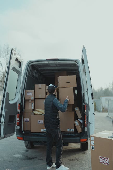 A man wearing a dark vest, dark pants, and a baseball cap stands inside the open rear of a large white moving van, which is parked outdoors on a paved surface. The van's interior is filled with multiple cardboard boxes of various sizes, some sealed with tape and marked with labels, stacked neatly to maximize space for a home relocation or furniture transport. One of the boxes is being lifted or positioned by the man, indicating an active loading process typical of packing and moving procedures. Several additional boxes are visible around the van's open doors, with some resting on the pavement nearby. The environment is outdoors with a cloudy sky and a background of trees and industrial structures, reflecting a commercial or residential moving context supported by the professional removals services of Man with Van Ilford. This detailed scene highlights the process of loading or unloading during a house removals operation, demonstrating careful handling of packing materials and organized furniture transport.