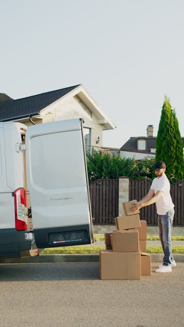 A man with a beard, wearing a white t-shirt and dark trousers, is standing outdoors next to an open white panel van on a residential street during daytime, with the back doors of the vehicle open. He