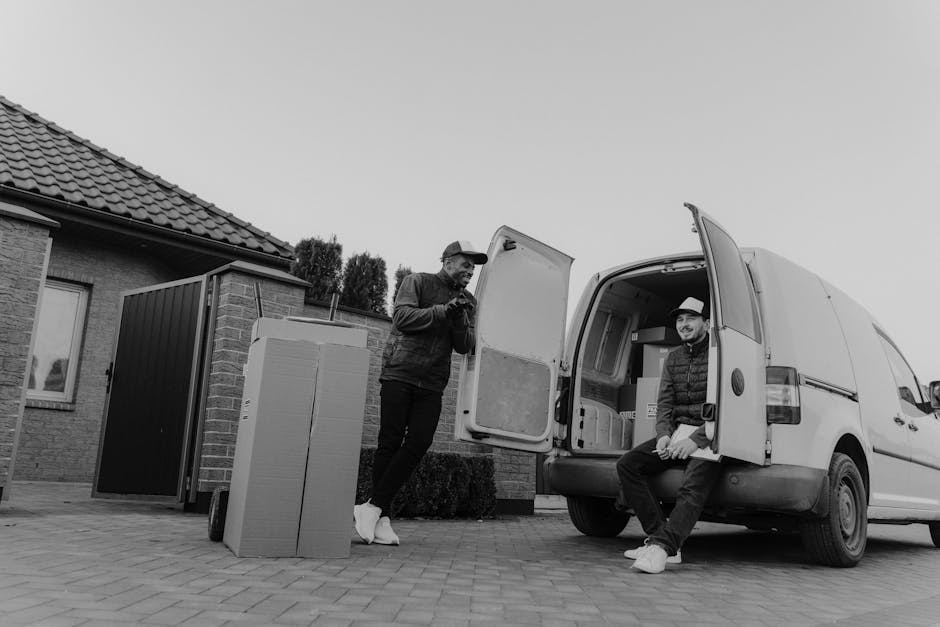 A man with a beard, wearing a white t-shirt and dark trousers, is standing outdoors next to an open white panel van on a residential street during daytime, with the back doors of the vehicle open. He