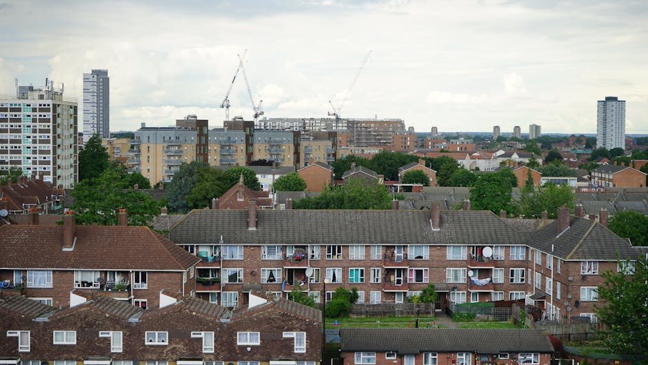 A panoramic view of a residential area featuring a row of three-storey brick apartment buildings with brown tiled roofs, situated in the foreground. Each apartment has small balconies with various items such as potted plants and satellite dishes visible on some. Behind these buildings, there is a mix of green trees and patchworks of rooftops of different shapes and materials, including flat concrete roofs and pitched tiled roofs. In the background, several high-rise construction cranes and modern apartment blocks under construction are visible, indicating ongoing development in the area. The sky above is overcast with a layer of grey clouds, creating diffuse natural lighting. The environment suggests an urban setting with potential logistical challenges for house removals, as elements like narrow pathways and limited access points are typical in such areas, which is relevant for services like those provided by Man with Van Ilford specializing in removals and furniture transport.