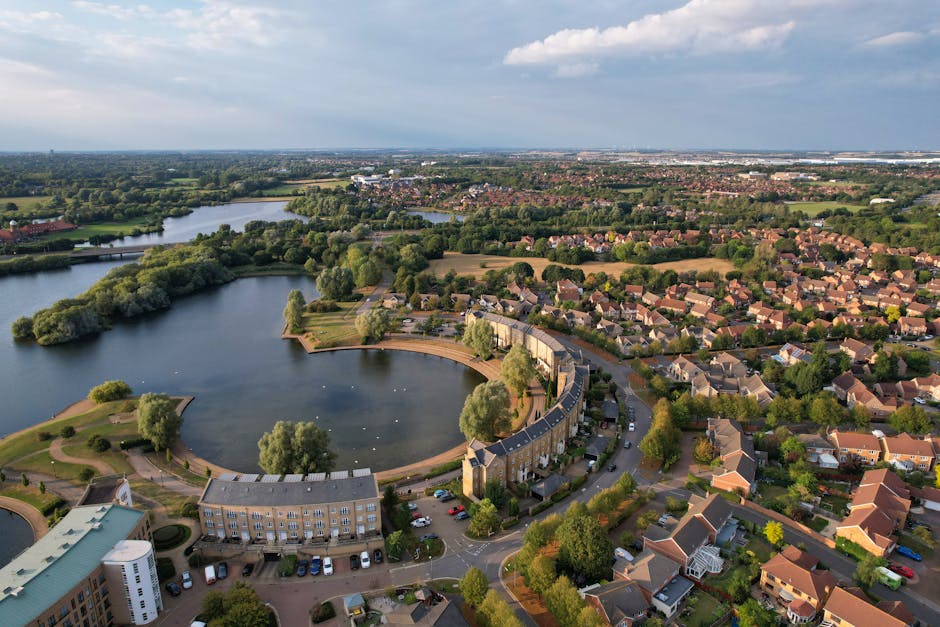 Aerial view of a residential area near a large, curved waterbody with a park and pathways along its edge. The water is calm with a few small boats, and the park features trees, grassy areas, and paved walkways. Surrounding the pond are rows of terraced houses, detached homes, and apartment buildings, with streets lined by trees and parked cars. In the background, additional green spaces, fields, and more distant buildings are visible under a partly cloudy sky. This scene relates to house removals and furniture transportation, illustrating the environment where Man with Van Ilford provides house relocation and packing services, with careful planning needed for accessing Cranbrook Estate for efficient moving and packing procedures.