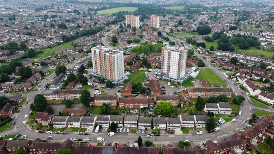 A panoramic view of a residential area featuring a row of three-storey brick apartment buildings with brown tiled roofs, situated in the foreground. Each apartment has small balconies with various items such as potted plants and satellite dishes visible on some. Behind these buildings, there is a mix of green trees and patchworks of rooftops of different shapes and materials, including flat concrete roofs and pitched tiled roofs. In the background, several high-rise construction cranes and modern apartment blocks under construction are visible, indicating ongoing development in the area. The sky above is overcast with a layer of grey clouds, creating diffuse natural lighting. The environment suggests an urban setting with potential logistical challenges for house removals, as elements like narrow pathways and limited access points are typical in such areas, which is relevant for services like those provided by Man with Van Ilford specializing in removals and furniture transport.