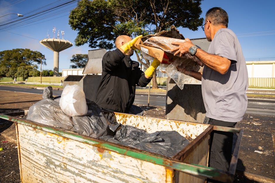 Two men are engaged in a house clearance or waste disposal activity outdoors, part of a home relocation process, with one man wearing a black jacket and yellow gloves lifting a cardboard box filled with assorted debris, including plastic packaging and other waste materials, while the other man in a grey T-shirt assists him by holding the box. They are positioned next to a large, rusty metal waste container loaded with black and grey garbage bags and additional waste. The scene takes place on a paved area near a street, with trees, a water tower, and a clear blue sky in the background. The environment suggests a waste collection or disposal site, possibly related to a removals or clearance service by Man with Van Ilford, involved in clearing household rubbish in preparation for moving or renovation, emphasizing the logistical process of loading and transporting waste as part of house clearance or home moving services.
