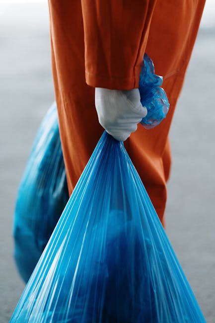 Close-up of a person dressed in an orange protective suit and white gloves, holding a large blue plastic garbage bag filled with waste material. The individual is standing outdoors on a paved surface, potentially near an entrance or driveway, with a slightly blurred background. The scene may relate to waste removal or cleaning services as part of a house removal or relocation process, illustrating the handling of waste disposal during moving activities, with the focus on procedures managed by Man with Van Ilford for efficient home clearance and compliance with waste disposal regulations.