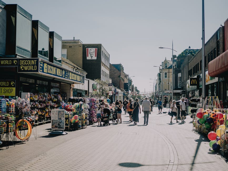 A busy high street in Ilford with various retail shops and outdoor stalls lining both sides of the pavement. Visible shopfronts include a discount store and a fast-food outlet, with merchandise such as colorful toys, balloons, and clothing displayed on racks and tables outside. Pedestrians of different ages walk along the street, some pushing strollers or carrying shopping bags, engaging in shopping or casual strolling. The street is illuminated by tall lampposts, and the scene is set during daytime with clear, bright weather and minimal shadows. The image captures a typical urban day involving foot traffic and outdoor shopping activities, relevant to home relocation or packing and moving services by Man with Van Ilford, especially in narrow street environments typical of Ilford High Road.
