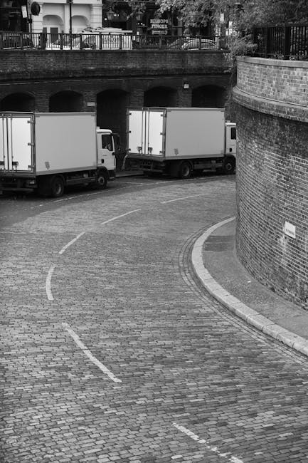 A narrow cobbled street in Ilford with a curved layout, showing three white box trucks parked along the side, some partially behind a brick wall and under a bridge structure with arches visible above. The trucks are positioned close to the pavement, which is lined with a low brick wall and a metal railing on the upper level. The scene appears to be during daylight with natural light, and the area looks suitable for home relocation and furniture transport involving careful navigation through tight streets, as would be relevant to house removals carried out by Man with Van Ilford. The image captures the logistical considerations of loading and unloading furniture on narrow streets, emphasizing the importance of planning for even small street access during a move.