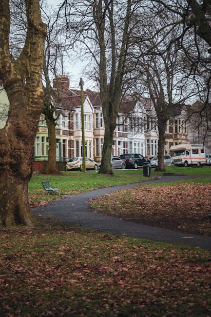 An outdoor scene in a residential neighbourhood with a curved paved footpath running through a small park area. The park has leafless trees with thick trunks and branches, and the ground is covered with fallen leaves. In the background, there are several traditional terraced houses with bay windows and brickwork, painted in various pastel colours such as light green and beige. Parked along the street in front of the houses are multiple cars, including a white van adjacent to a driveway. A bench is situated near the pathway, and a black trash bin is visible near one of the houses. This setting suggests preparation for a house removal, with ample space for furniture transport and loading, and is indicative of a typical residential street in Ilford. The presence of large trees and parked vehicles highlights the logistical considerations involved in home relocation and moving services provided by Man with Van Ilford.