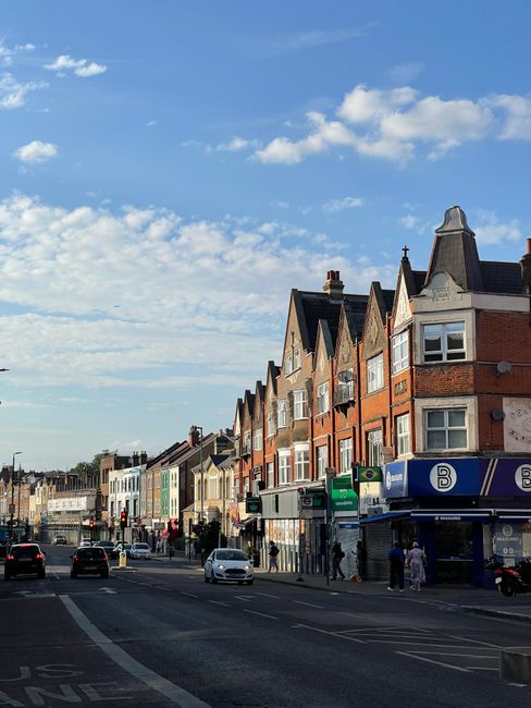 This image shows a typical residential street in Ilford with a row of multi-storey houses featuring red brick facades, bay windows, and pitched roofs, some with chimney stacks. The street is busy with