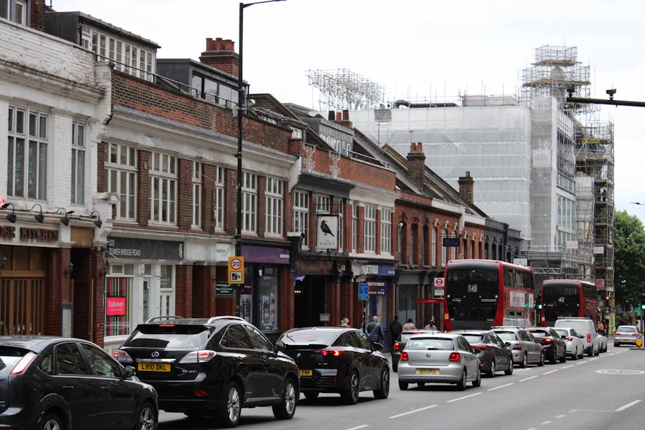 A busy street scene in Ilford during daytime showing a row of brick buildings with shopfronts, some under renovation surrounded by scaffolding and protective sheeting. In the foreground, a line of parked cars and SUVs, including a black vehicle with a roof rack, extends along the pavement. Pedestrians are visible walking on the sidewalk near the storefronts, some carrying bags or packages. A red double-decker bus is actively moving along the street, with route signage visible. The street is equipped with signage indicating parking restrictions and speed limits. The scene is well-lit with natural daylight, highlighting the urban environment and ongoing building work, which suggests preparation for a home relocation or furniture transport process managed by Man with Van Ilford, consistent with house removals and moving services.