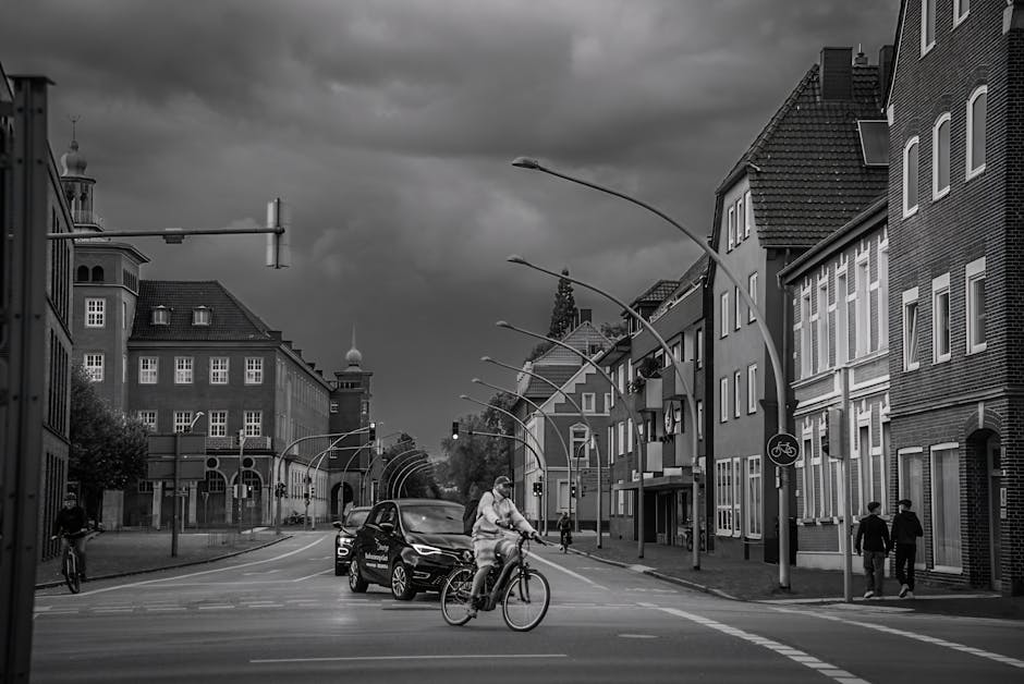 A black and white photo of a city street scene during daytime, showing a cyclist riding across an intersection in the foreground, with a woman pushing a small trolley containing cardboard boxes nearby. A black van is stationary further along the road, likely involved in a home relocation or moving process, with its side door open next to a building's doorway. Several multi-story residential buildings with brick facades and large windows line both sides of the street, some with small balconies, and a few pedestrians are walking along the pavement. The street is equipped with street lamps, traffic signals, and a cyclist path sign, indicating facilities for both pedestrians and cyclists. Overhead, curved street lights are mounted on tall poles, and dark, cloudy skies suggest an overcast day. This scene illustrates typical urban logistics involved in furniture transport and packing during a home move, captured near Ilford Station, where [COMPANY_NAME] occasionally facilitates removals and relocation services, ensuring the safe loading and transport of household items from residential buildings to vehicles.