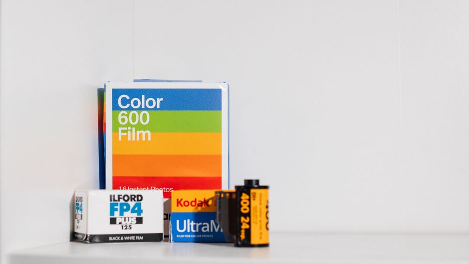 A collection of photographic materials and equipment are arranged on a white surface inside a property, preparing for home relocation or furniture transport. The arrangement includes a box of Ilford FP4 Plus black and white film, positioned behind a smaller box of Kodak Ultra Max film, signifying packing for camera equipment. To the left, there is a packet of Color 600 Film, partially propped against the other boxes, with vibrant rainbow stripes on its packaging. In front, a pack of 35mm black and white film is visible, along with a roll of 35mm film labeled '400-24', both indicating preparation for photographic media packing. The items are situated near an open doorway or window, with natural light illuminating the scene, emphasizing careful packing of fragile items typical during moving or packing processes. The setting suggests ongoing or upcoming relocation services provided by Man with Van Ilford, focusing on professional packing, loading, and transportation of personal belongings, including delicate and valuable photographic equipment.